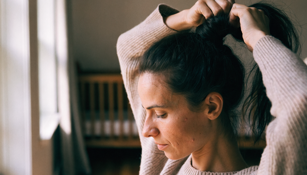 Jeune mère attachant ses cheveux près d'une fenêtre — chute de cheveux post-partum